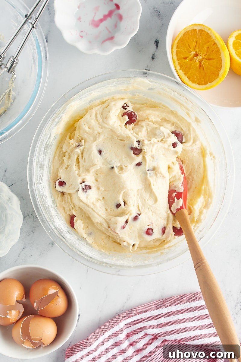 Gently folding in cranberries into the Bundt cake batter with a spatula to ensure even distribution without crushing.