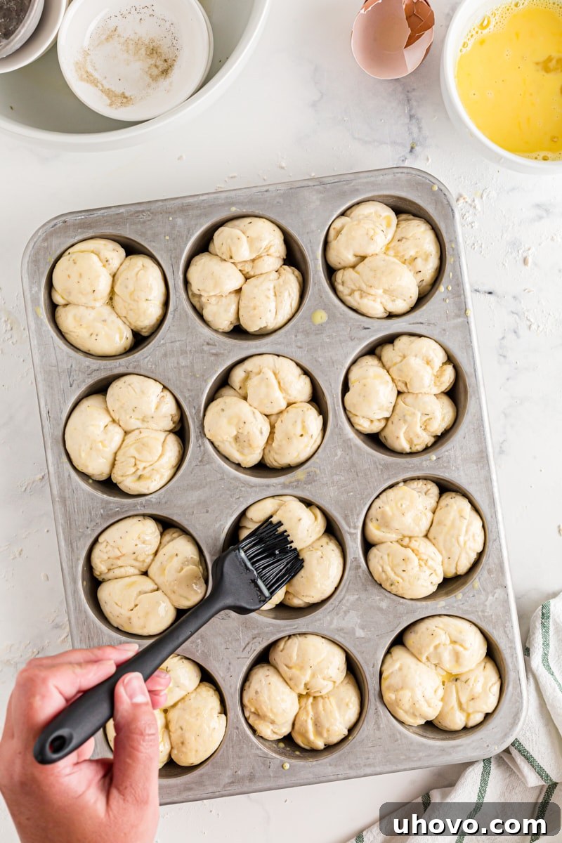 A pastry brush applying a glistening egg wash over the risen dough balls for Herb Cloverleaf Rolls in a muffin tin.