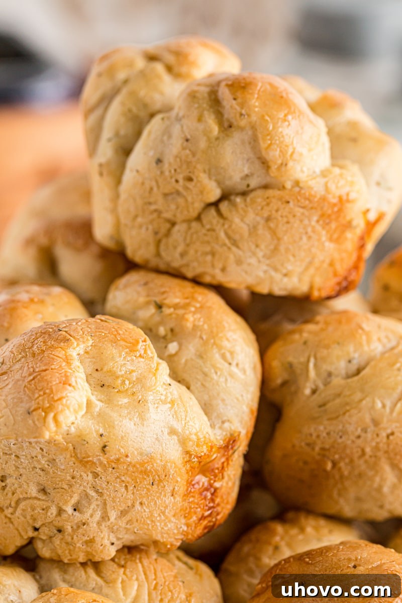 A close-up of a pile of golden-brown cloverleaf dinner rolls, with visible flecks of green herbs baked into their fluffy dough.