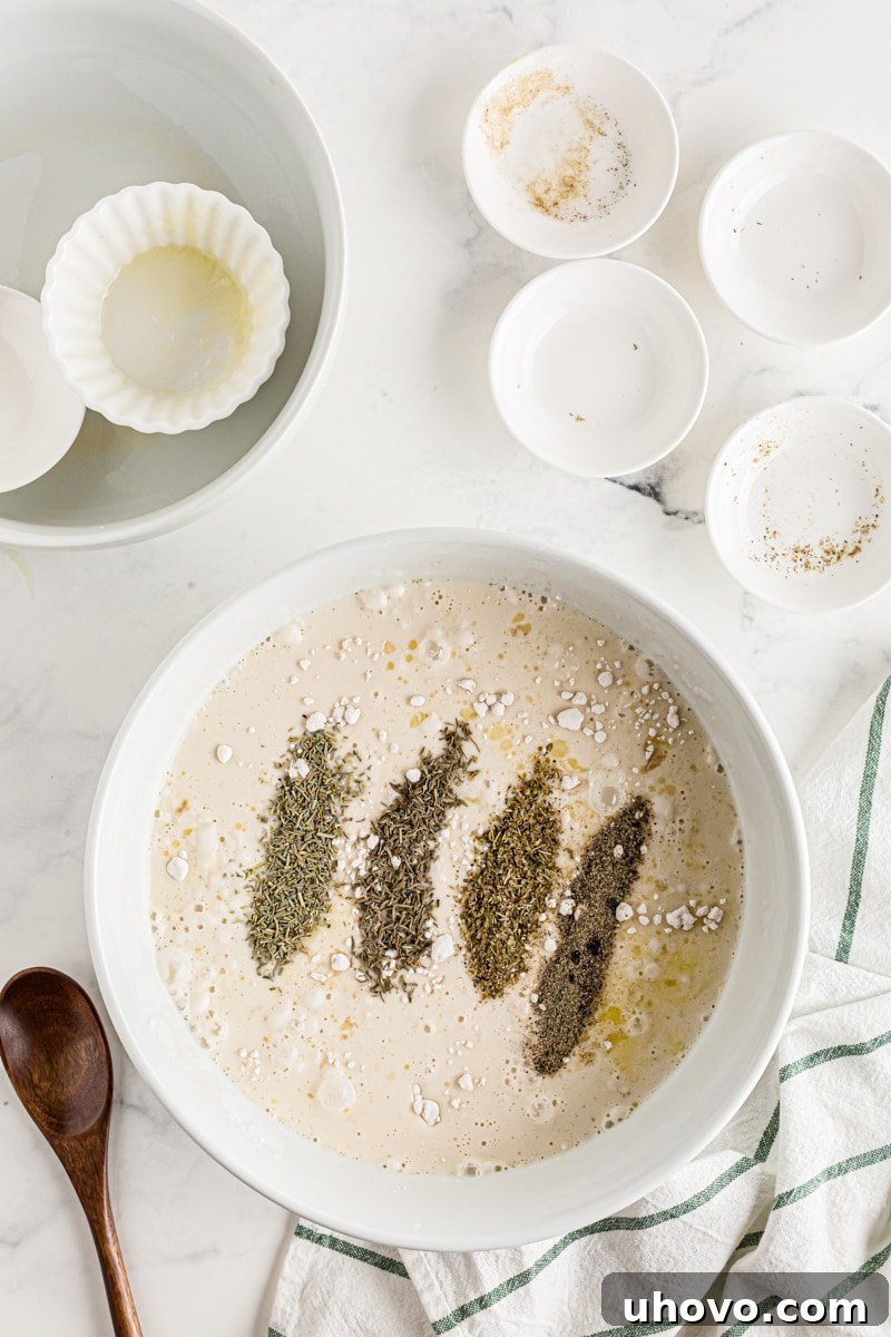 Dried herbs and spices being sprinkled over the soft dinner roll dough in a mixing bowl.