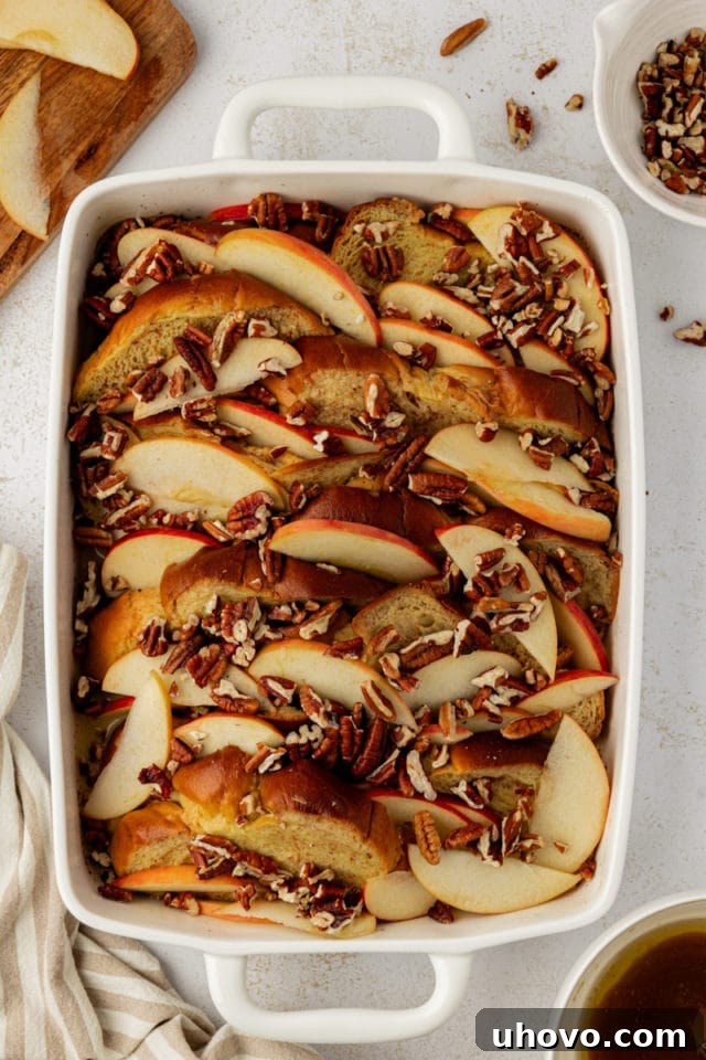 Chilled apple cinnamon baked French toast in a baking dish, topped with apple slices and pecans, awaiting the final butter-sugar drizzle before baking.