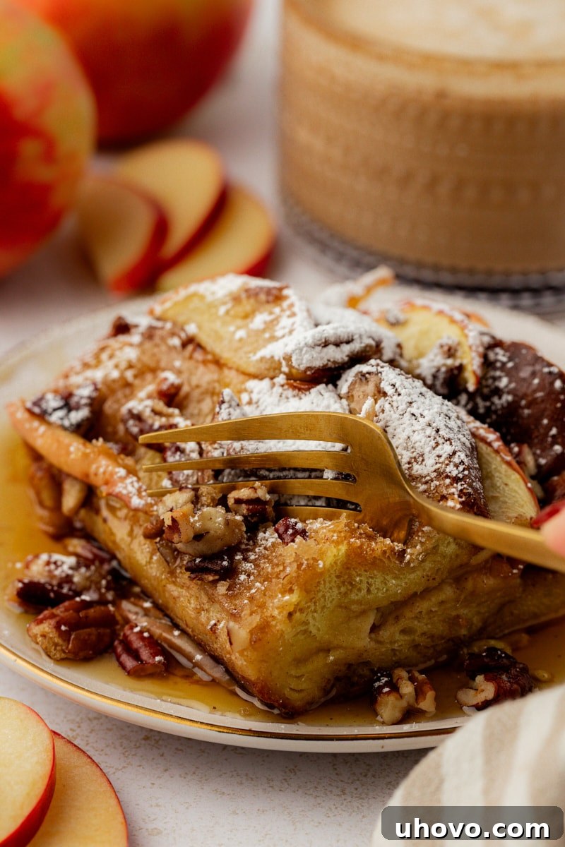 A fork gently cuts into a corner slice of apple cinnamon French toast, revealing its soft, custardy interior with visible apple pieces and pecans.