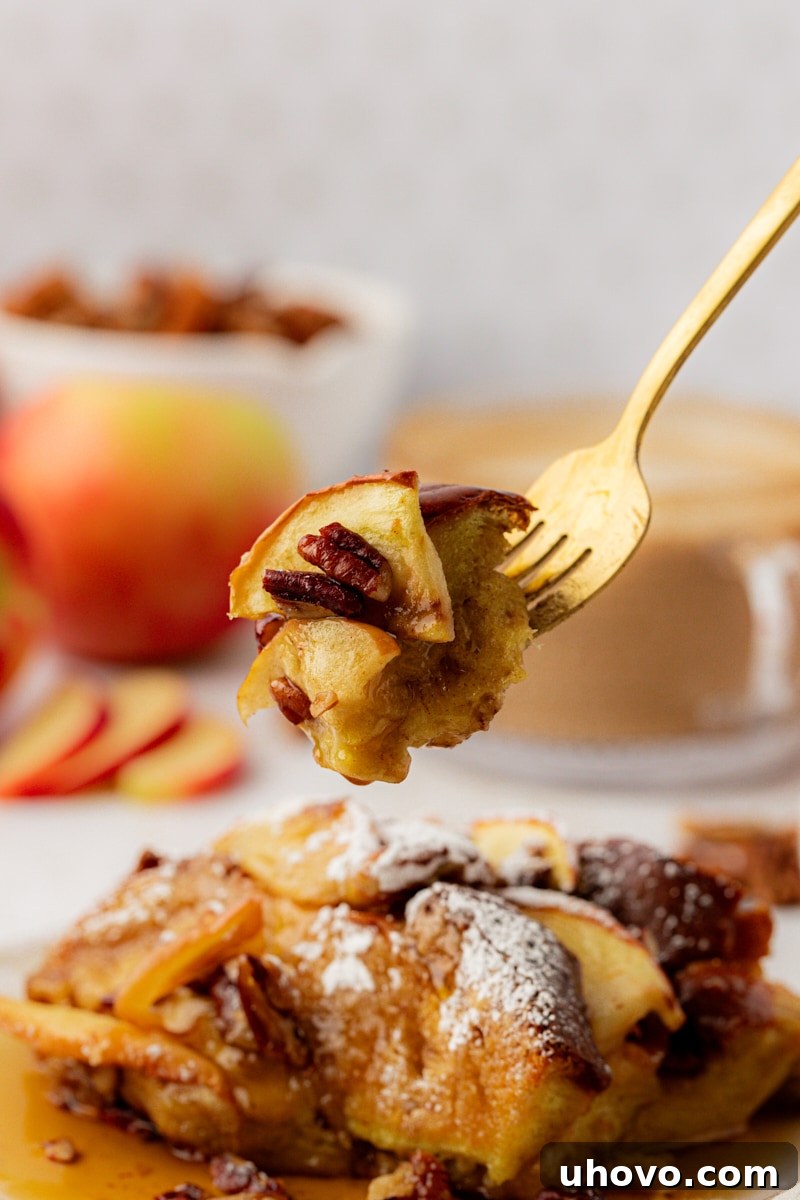 A fork holds a perfect bite of apple cinnamon French toast, showcasing the tender bread, apple chunks, and pecans, poised above a plate with more slices.