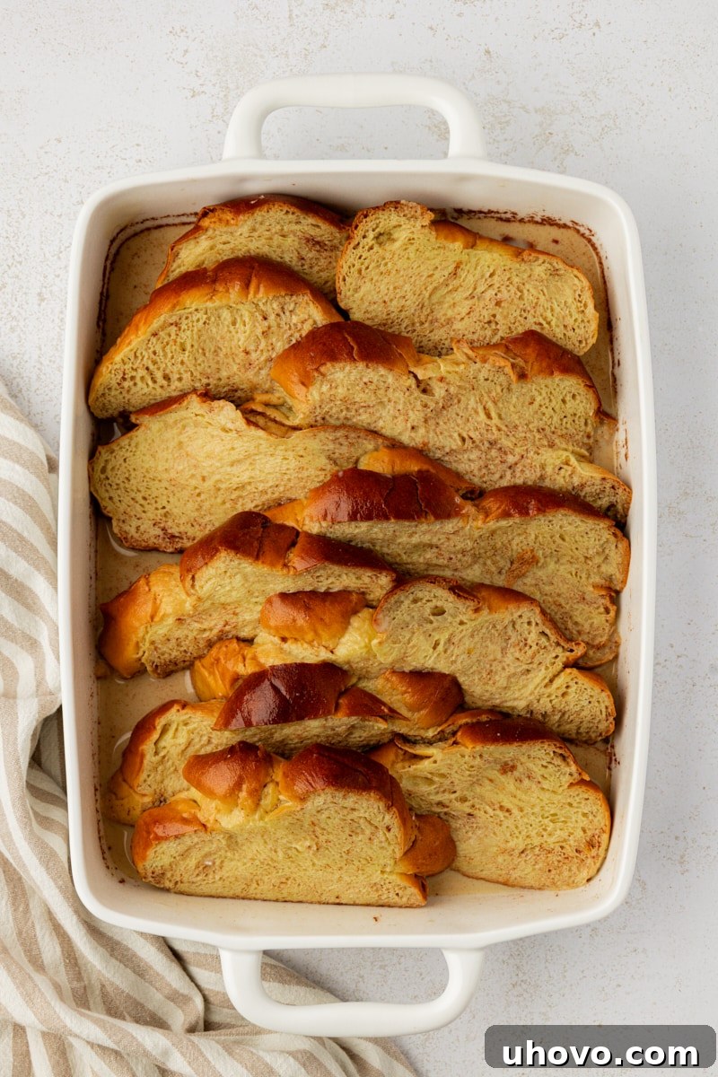 Overhead view of challah bread slices submerged in a rich, spiced custard within a baking dish, ready for chilling.