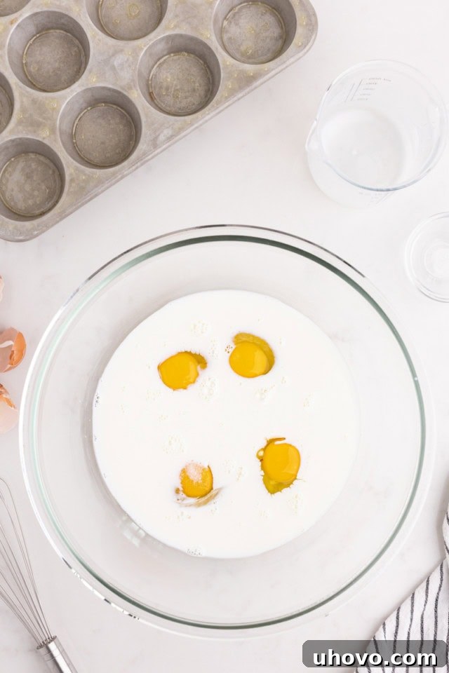Overhead view of eggs, warm milk, and salt being whisked together in a clear glass mixing bowl.