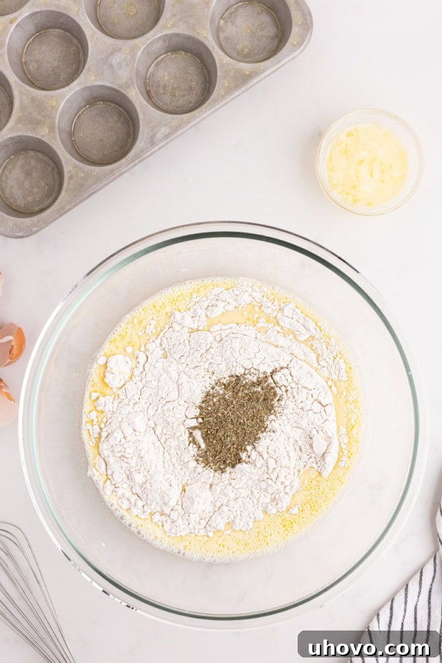 Overhead view of all-purpose flour and fresh herbs being added to the wet popover batter in a glass mixing bowl.