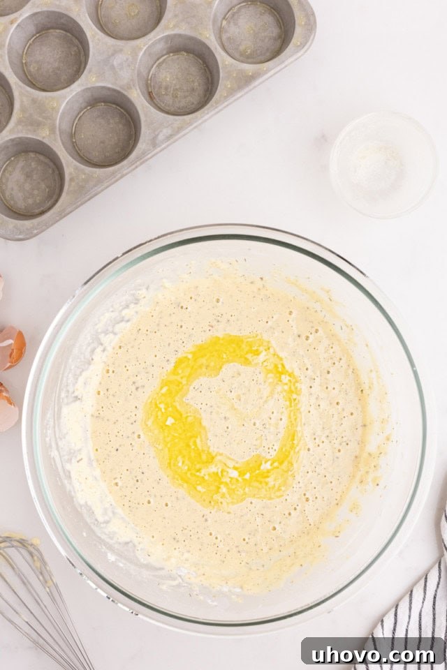 Overhead view of melted unsalted butter being gently stirred into the frothy popover batter in a glass mixing bowl.