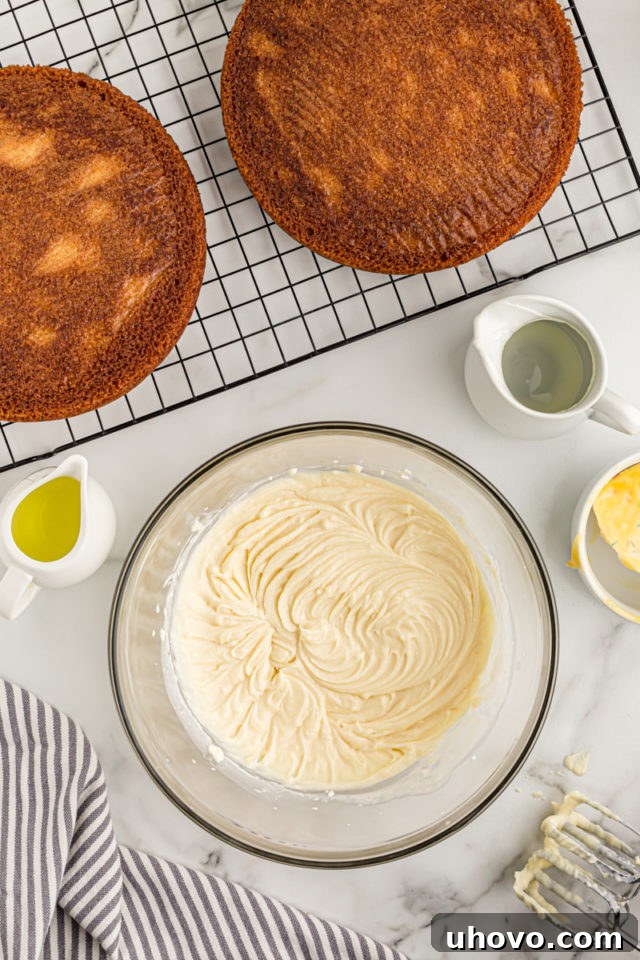 Overhead view of the finished mascarpone frosting in a glass mixing bowl, with cooled cake layers visible on a wire rack in the background.