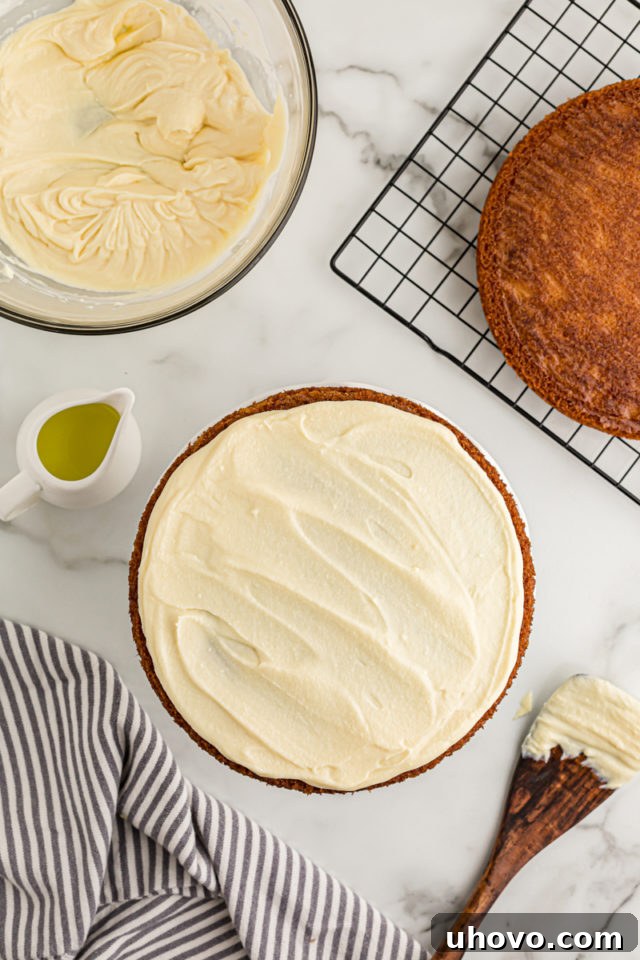 Overhead view of the first frosted lemon cream cake layer, with the second unfrosted layer waiting on a rack in the background.
