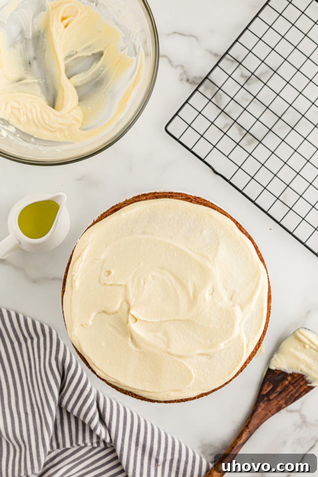 Overhead view of the completely frosted lemon cream cake, showcasing its smooth finish, with a wire rack, bowl of frosting, and spatula in the blurred background.