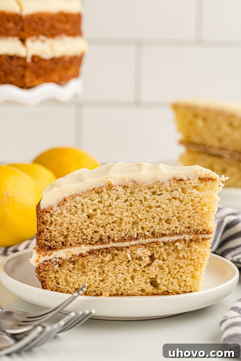 Side view of a gorgeous lemon cream cake, beautifully layered and frosted, presented on a white cake plate. Another cake is blurred in the background.
