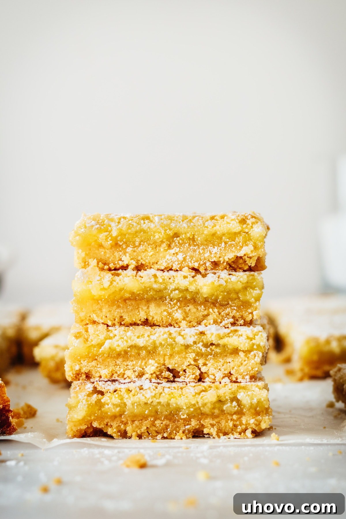 A stack of four perfectly cut lemon bars, glistening with powdered sugar, resting on parchment paper. Each bar shows off its golden crust and bright yellow filling.