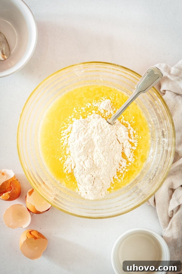 Overhead view as the dry flour and baking powder mixture is gently folded into the wet lemon, egg, and sugar mixture in a large mixing bowl.