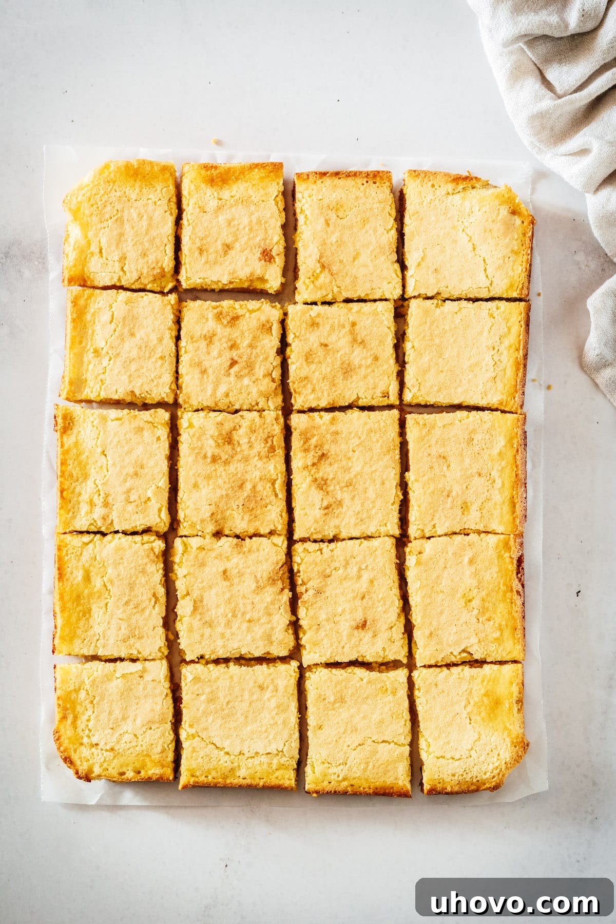 Overhead view of a tray of freshly baked lemon bars, cooled and neatly cut into squares on parchment paper, before the final dusting of powdered sugar.