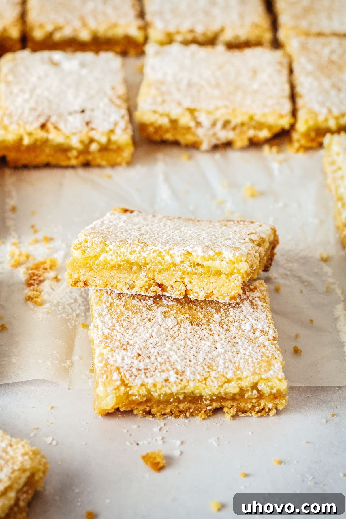 Two perfectly cut lemon bar squares, one stacked on top of the other, with more delicious bars visible in the softly blurred background.