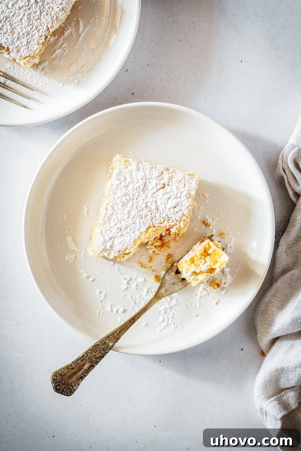 A single lemon bar resting on a white plate with a fork, a small bite already taken, highlighting the soft filling and crumbly crust.