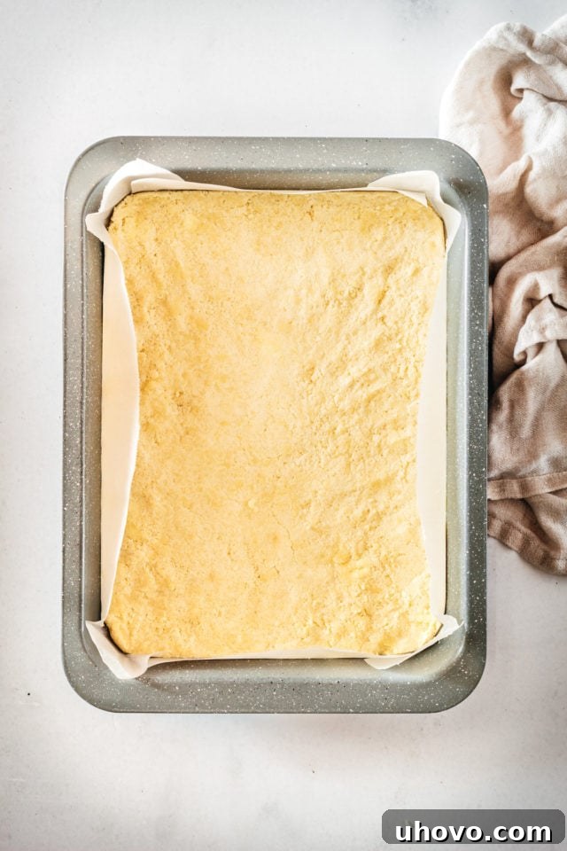 Overhead view of the shortbread crust mixture pressed evenly into a parchment-lined baking pan, ready for its initial bake.