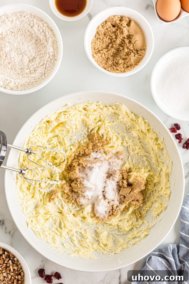 An overhead shot shows golden brown and granulated sugars being carefully added to creamed unsalted butter in a pristine white mixing bowl, ready for the next step in cookie dough preparation.