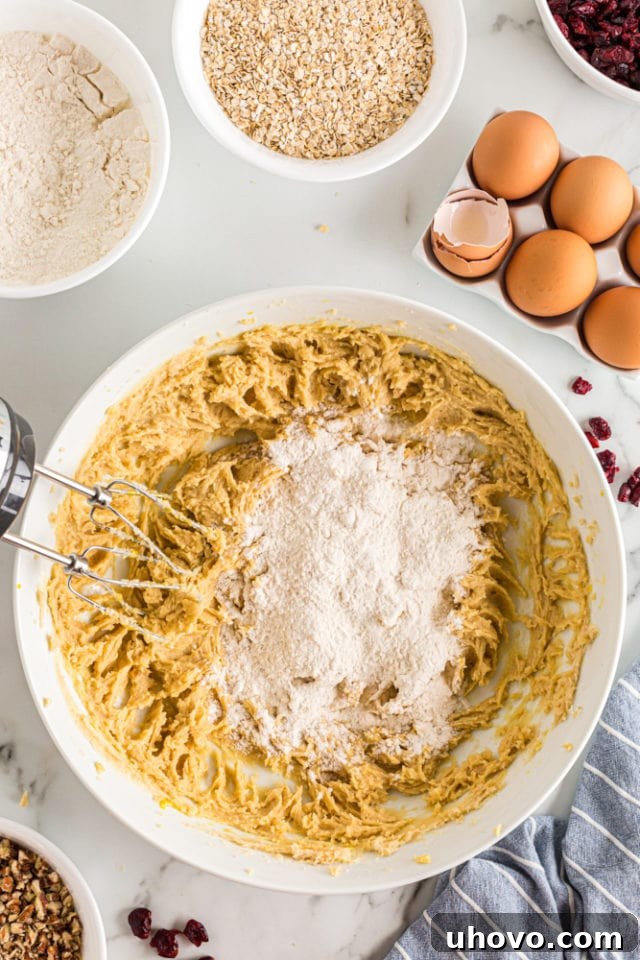 An overhead view captures the moment dry flour mixture is being slowly added in small portions to the wet ingredients in a white mixing bowl, ensuring gradual incorporation for optimal cookie dough consistency.