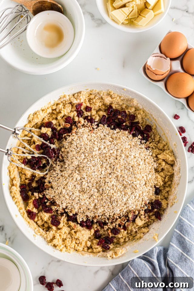 An overhead shot captures the vibrant addition of dried cranberries, crunchy pecans, and wholesome quick oats being sprinkled into the creamy cookie dough in a white mixing bowl, ready for gentle folding.