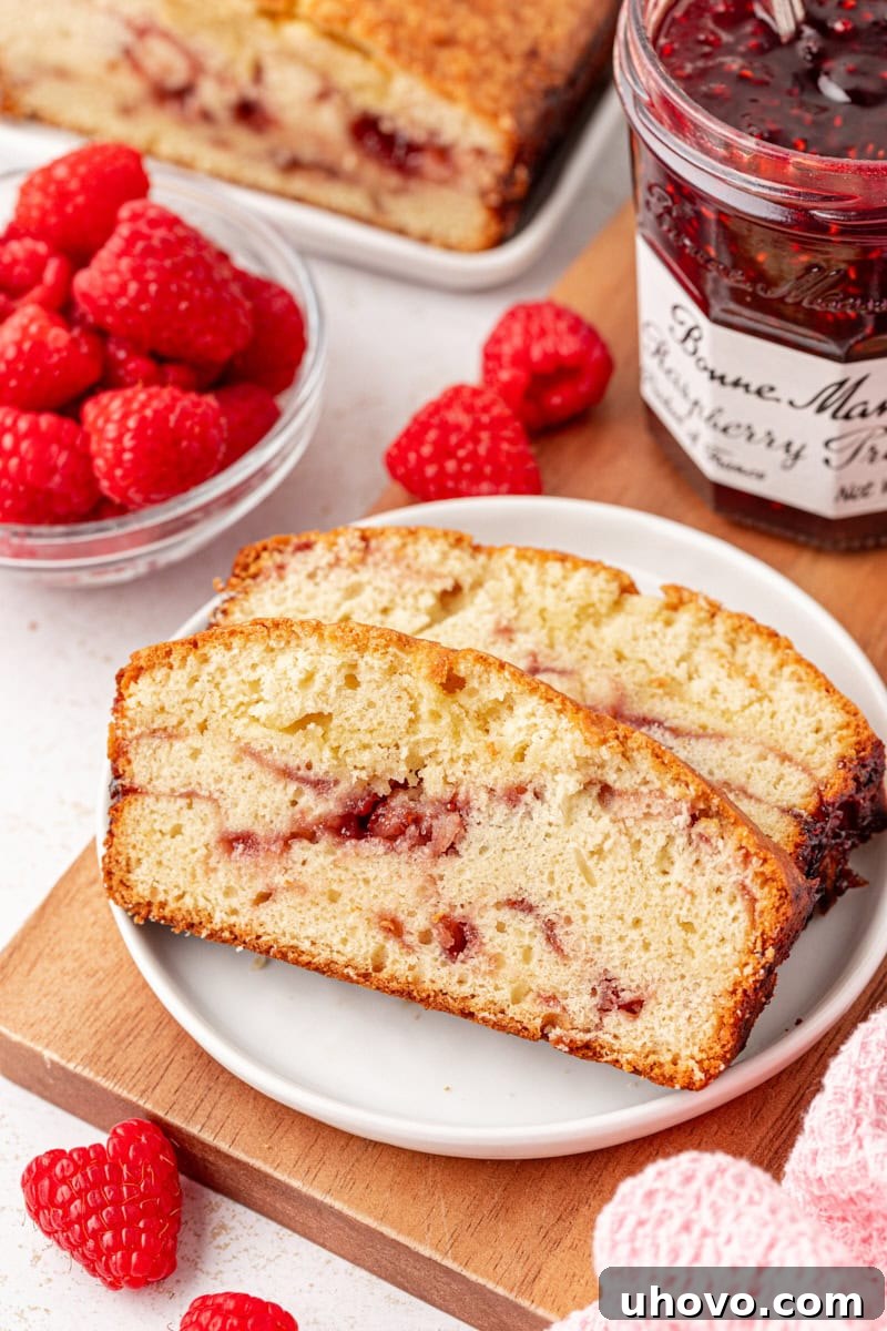 Two slices of berry swirl pound cake on a white plate, surrounded by fresh raspberries, more cake, and a jar of raspberry preserves. The cake is soft, buttery, and beautifully swirled with fruit.