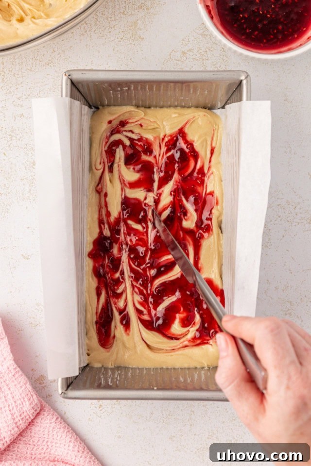 Overhead view of berry preserves being swirled into cake batter in a loaf pan, showing distinct layers.