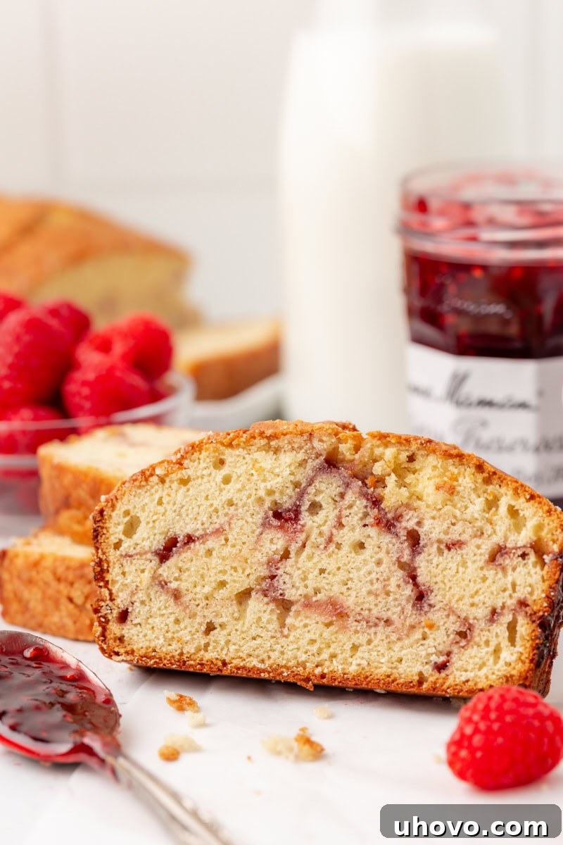 A slice of berry swirl pound cake on a white plate, with more cake, fresh raspberries, and a jar of raspberry preserves in the soft-focus background, creating an inviting scene.