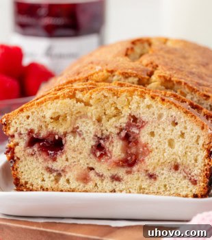 Partially sliced berry swirl pound cake on a white plate with fresh raspberries and a jar of raspberry preserves in the background, ready to be enjoyed.