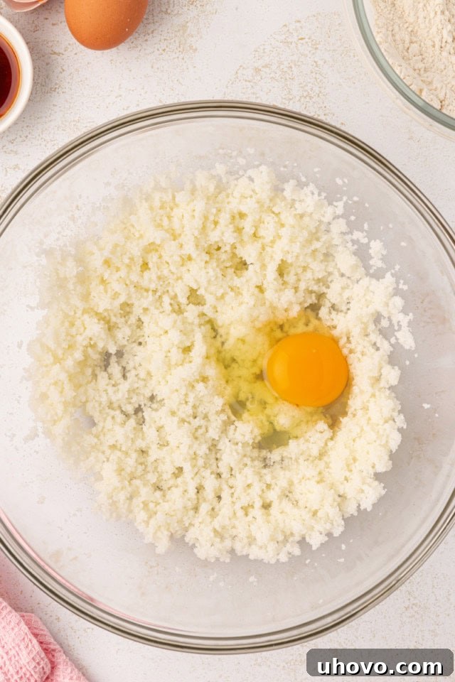 Overhead view of an egg being added to creamed butter and sugar in a glass mixing bowl, along with vanilla extract.