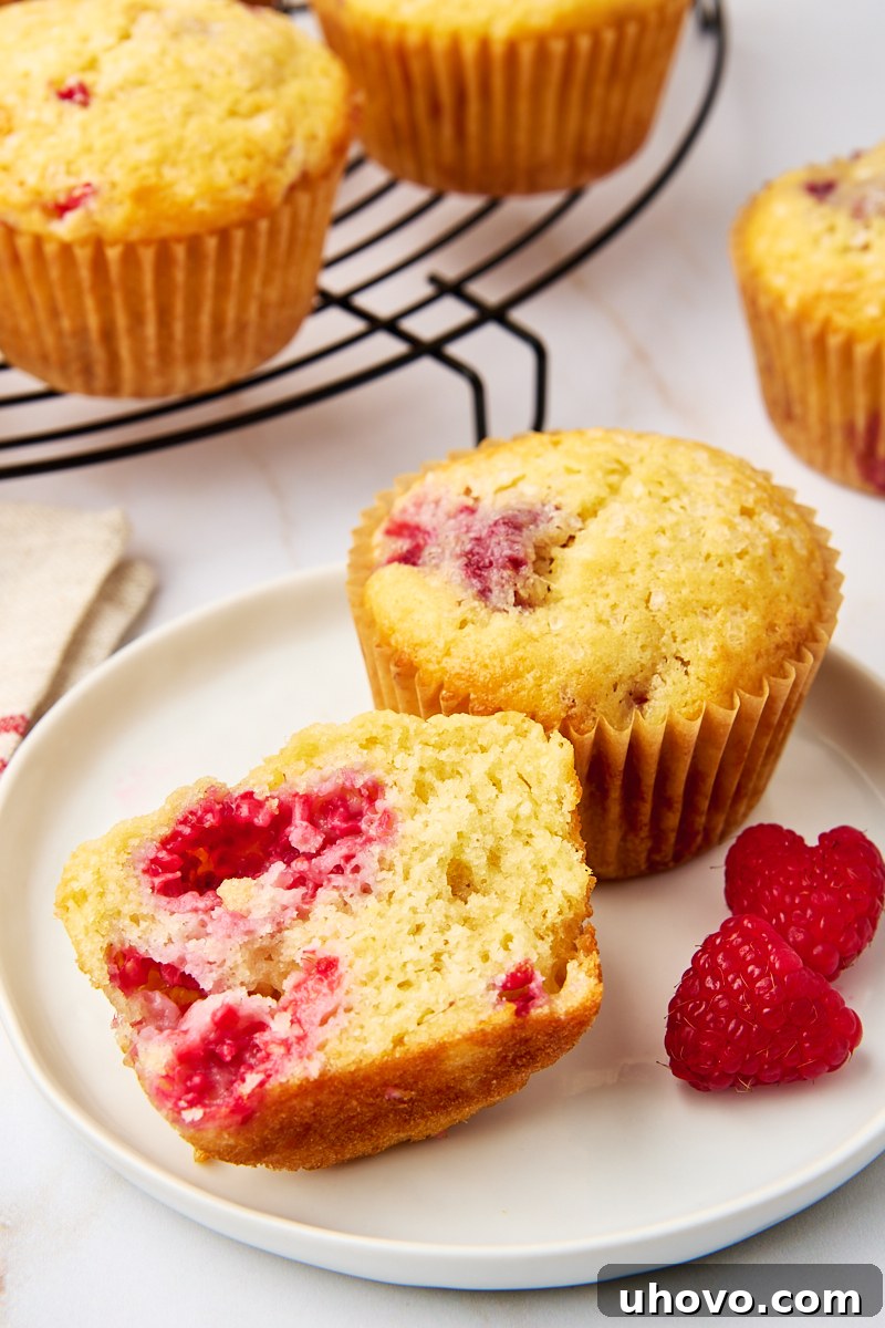 A close-up shot of a lemon raspberry muffin cut in half, revealing its tender crumb and juicy raspberries, with another whole muffin on a white plate in the background.