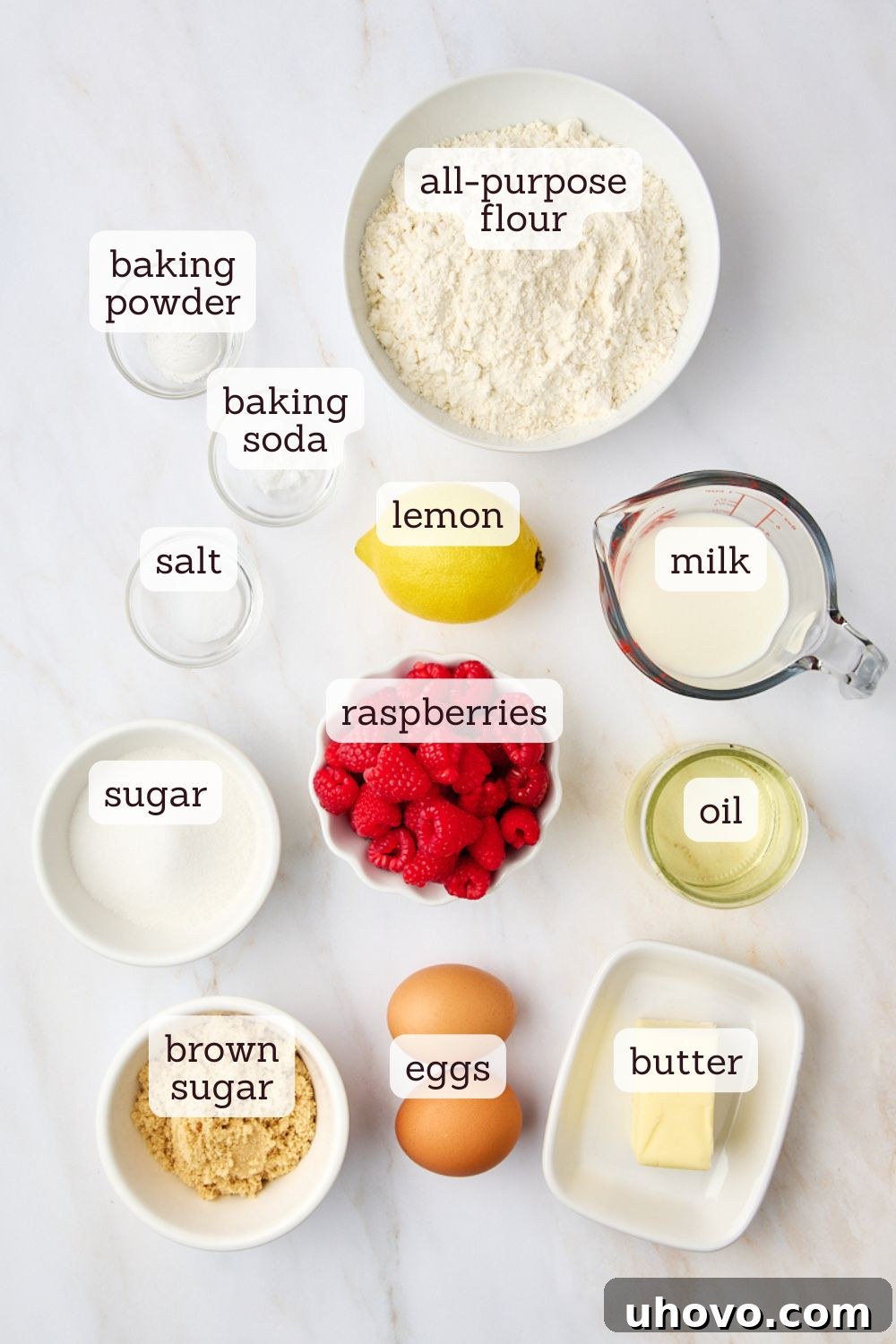 An overhead view of various raw ingredients neatly arranged on a white surface, including flour, sugars, butter, eggs, milk, fresh lemons, and raspberries, ready for making muffins.