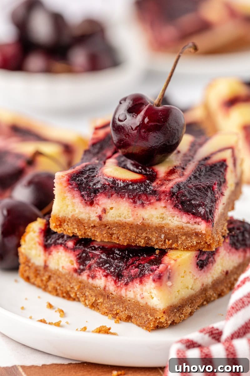 A stack of two perfectly sliced cherry cheesecake bars, garnished with a fresh, ripe cherry on top, highlighting the vibrant red swirl against the creamy white cheesecake and golden graham cracker crust.