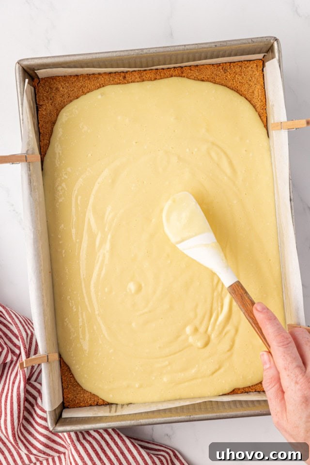 A visual of the smooth cheesecake batter being evenly spread over the partially baked graham cracker crust in the baking pan, creating a pristine layer.