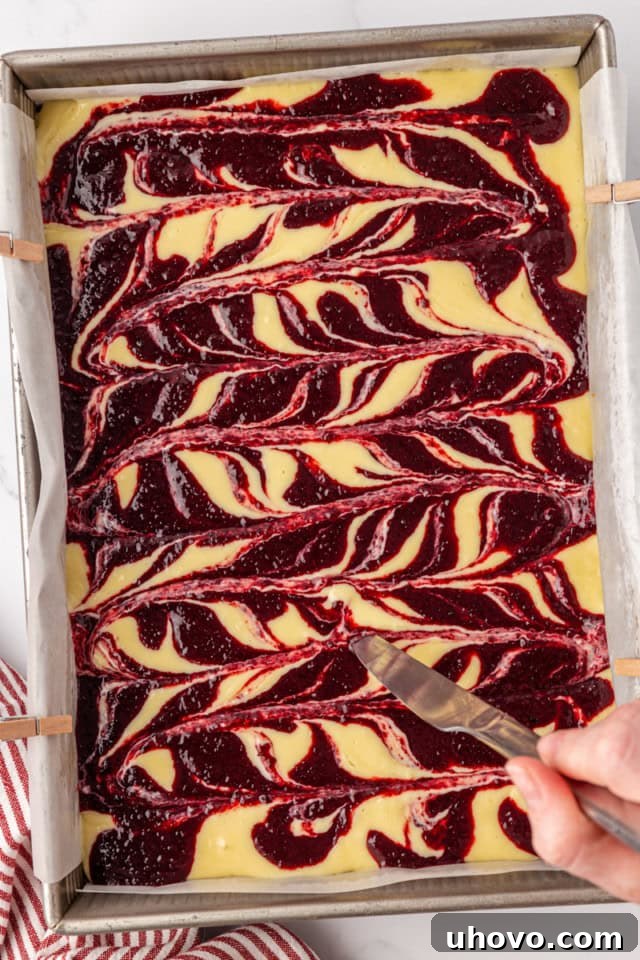 A thin knife gently swirls the vibrant cherry topping into the creamy cheesecake filling, creating an artistic marbled pattern on the surface before baking.