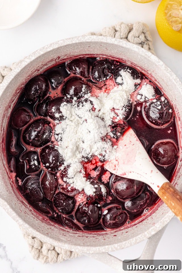 A close-up, overhead view of partially cooked cherries in a saucepan, with cornstarch and a squeeze of fresh lemon juice being added to the mixture, ready to be stirred in for thickening.