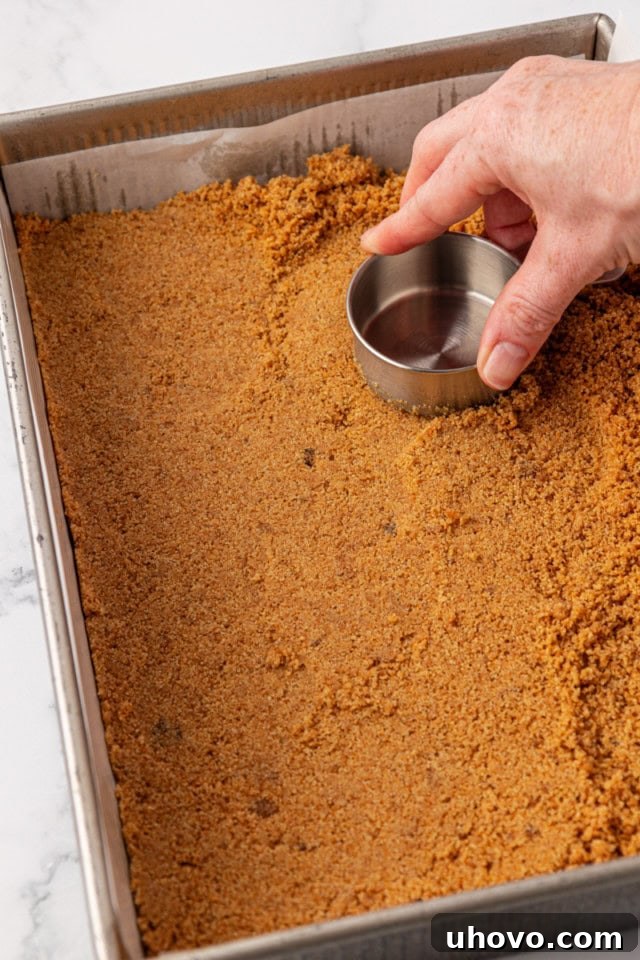 A visual of the graham cracker crust being firmly and evenly pressed into the bottom of a 9x13 inch baking pan, creating a compact base for the cheesecake bars.