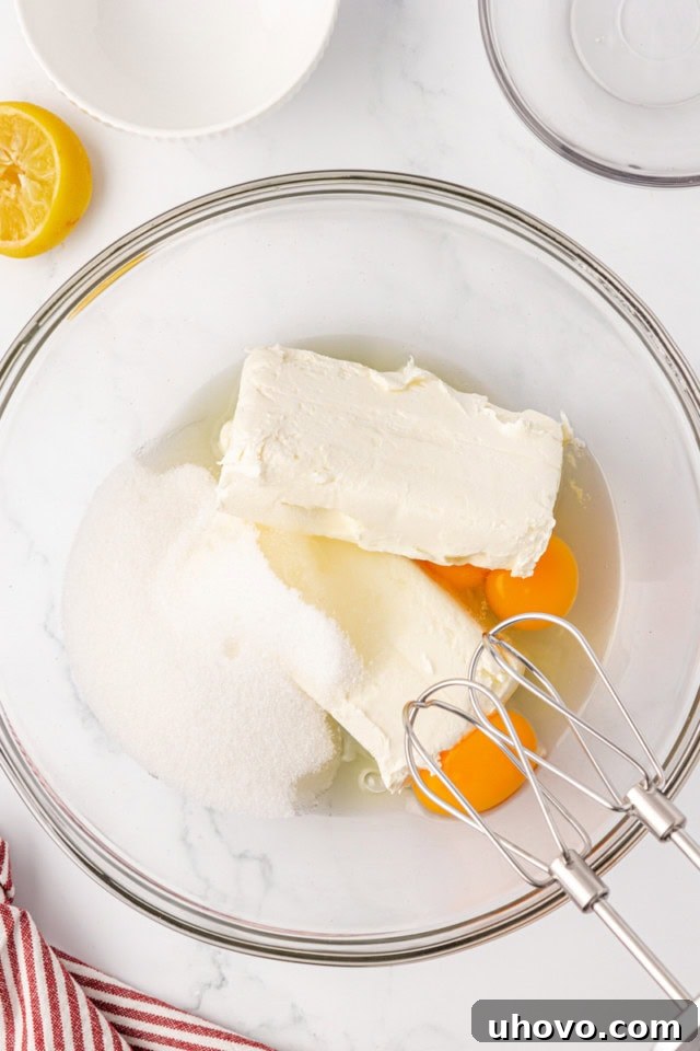 An overhead view of cream cheese, sugar, eggs, and fresh lemon juice gathered in a large glass mixing bowl, ready to be beaten into a smooth cheesecake filling.