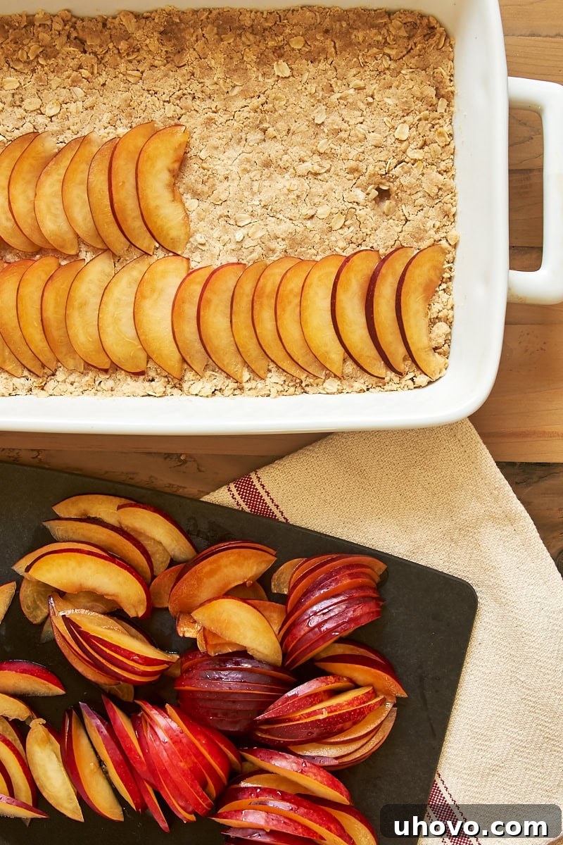 An overhead view of partially assembled plum crumble bars in a white baking dish, with freshly sliced plums ready on a wooden cutting board beside it, illustrating the preparation process.