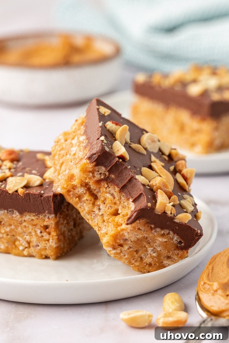 A single chocolate peanut butter rice crispy treat with a small bite taken out, resting next to another full treat on a white plate, highlighting its tempting layers.
