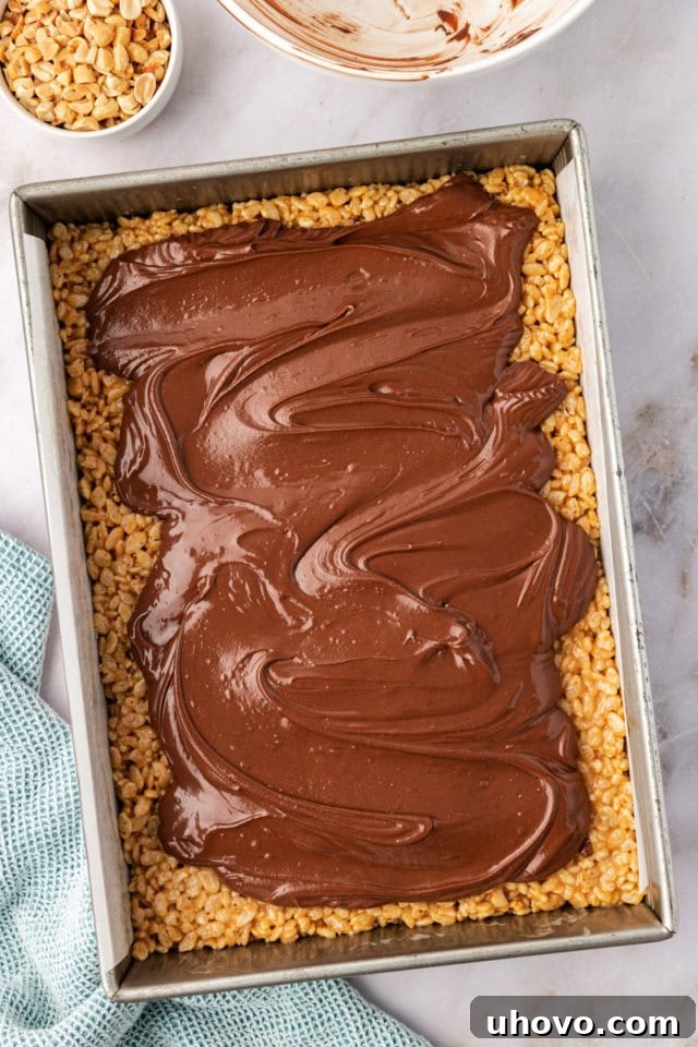 A partially spread layer of melted chocolate and peanut butter chips elegantly covering the crispy treat base, with roasted peanuts beginning to be sprinkled on top.