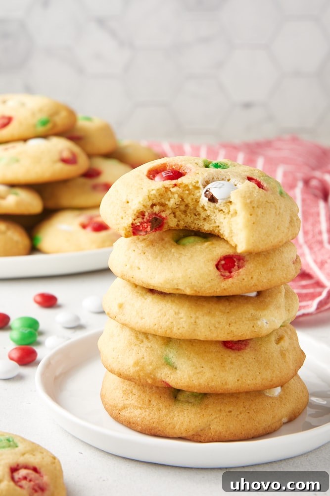 5 Christmas M&M cookies stacked on a white plate with a bite missing from the top one.