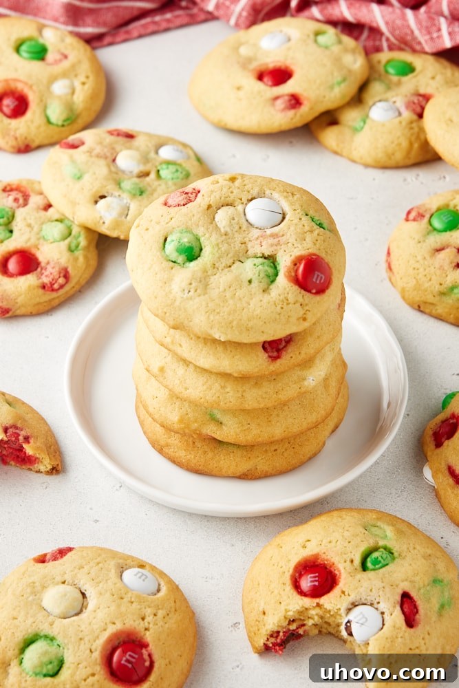 M&M cookies stacked on a white plate with more cookies surrounding.
