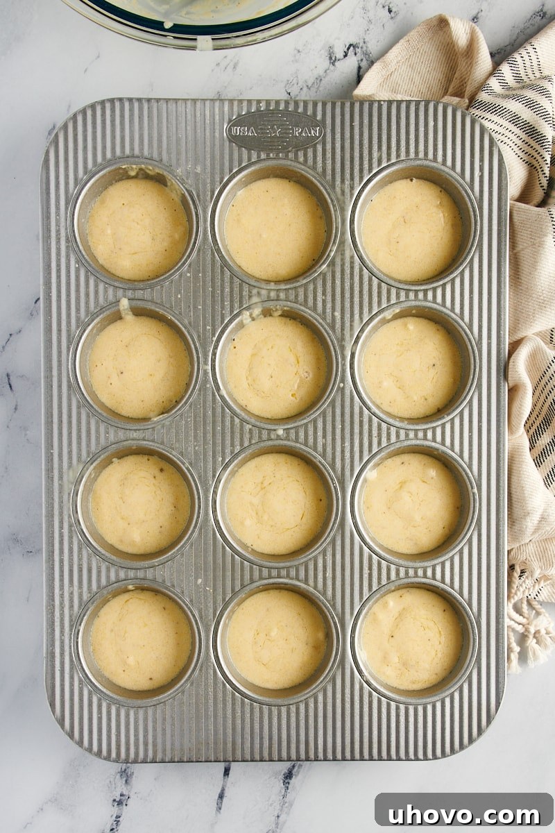 A standard 12-cup muffin tin, with each cup neatly filled about three-quarters full with smooth, golden corn muffin batter.