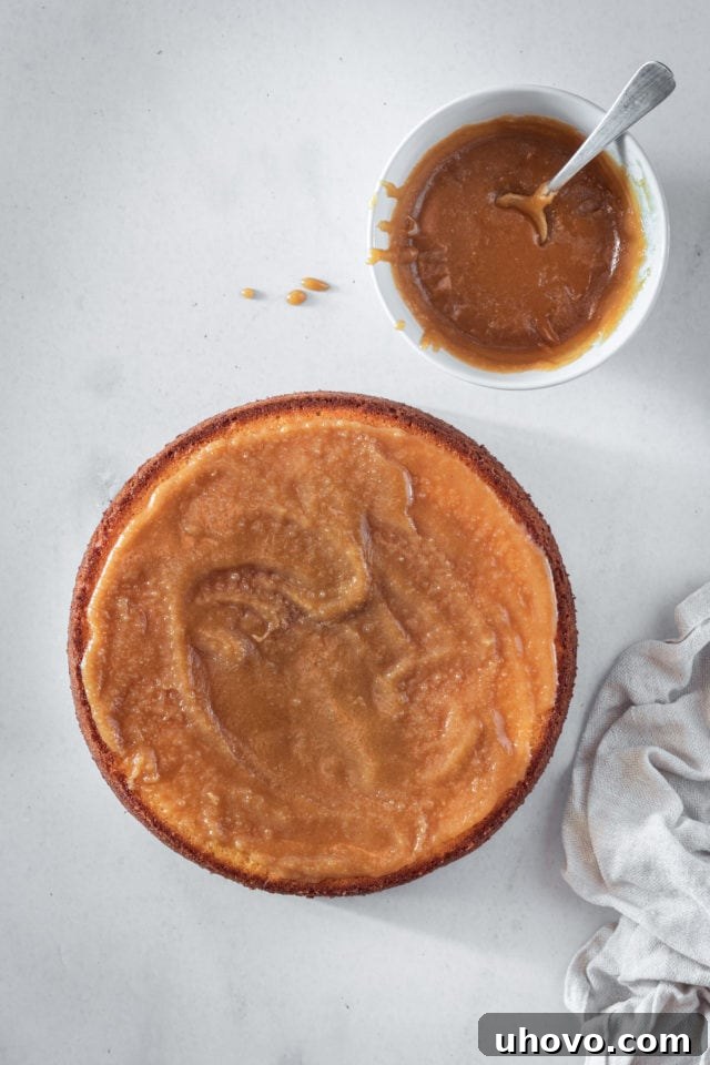 An overhead shot of warm, glistening caramel being spread evenly across a baked layer of cake, demonstrating the filling of the Southern caramel cake before the next layer is added.