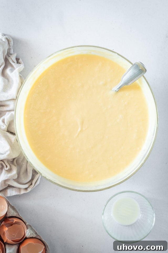 An overhead shot showing the smooth, golden caramel cake batter in a clear glass mixing bowl, ready to be divided into cake pans, illustrating the final mixing stage.