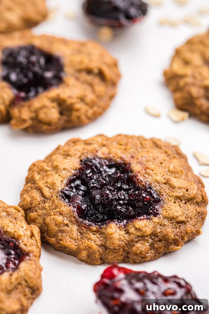 closeup of an oatmeal thumbprint cookie filled with blackberry jam on a white surface with more cookies in the background
