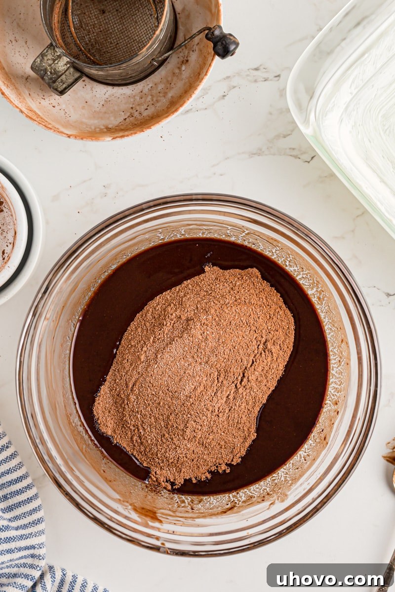 Overhead view of granulated sugar and unsweetened cocoa powder being added to the melted chocolate and butter mixture in a bowl.