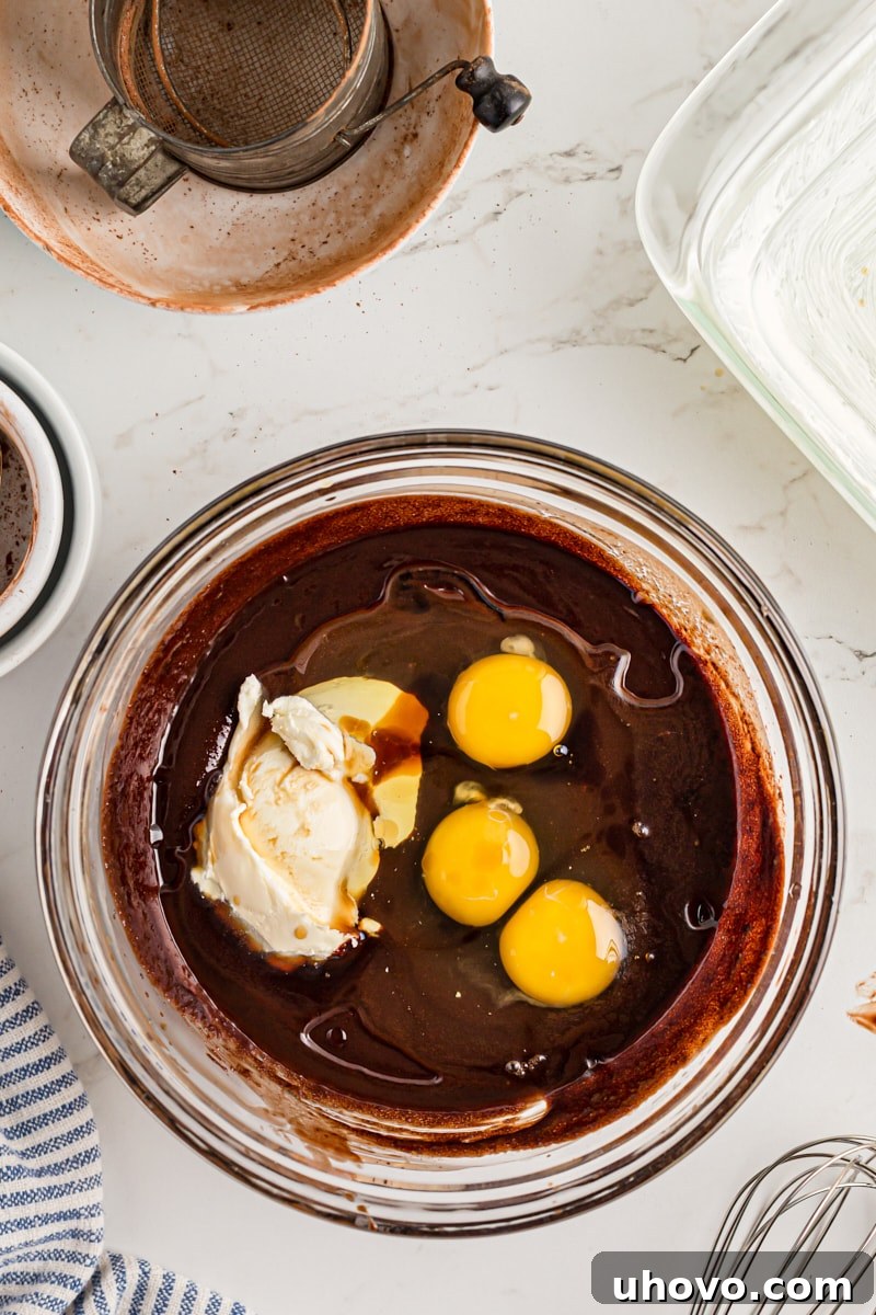 Overhead view of softened mascarpone cheese and eggs being added to the wet brownie ingredients in a mixing bowl.