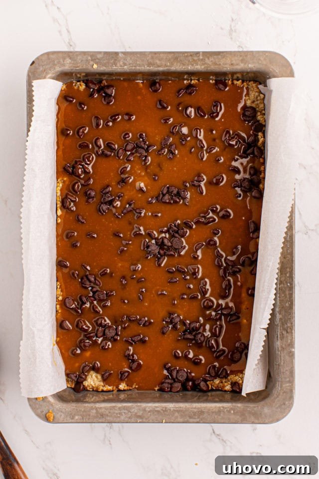 Warm caramel mixture being poured evenly over the chocolate chips and oat crust.