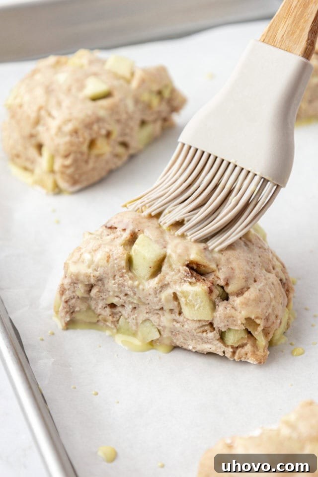 A hand brushing egg wash onto uncooked apple cinnamon scones arranged on a baking sheet.